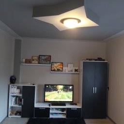 Remonty i wykończenia Marcin Dydak - Jasne wnętrze pokoju z szarą ścianą, telewizorem i lampą w kształcie gwiazdy na suficie. Widoczne meble w odcieniach bieli i czerni oraz ciemny dywan.