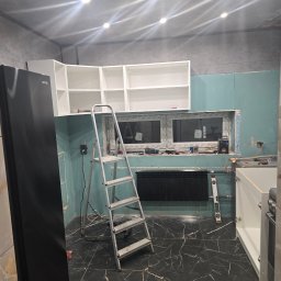 Remonty i wykończenia Marcin Dydak - Kuchnia w trakcie remontu: zamontowane białe szafki wiszące, ściany z płyt gipsowo-kartonowych, drabina aluminiowa, czarna marmurowa podłoga i widoczne narzędzia.