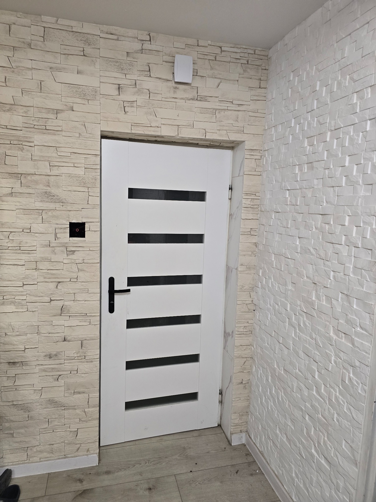 Białe drzwi z czarną klamką w Wojkowicach Kościelnych, wkomponowane w ścianę wyłożoną dekoracyjną płytką imitującą kamień. Nowoczesny design.