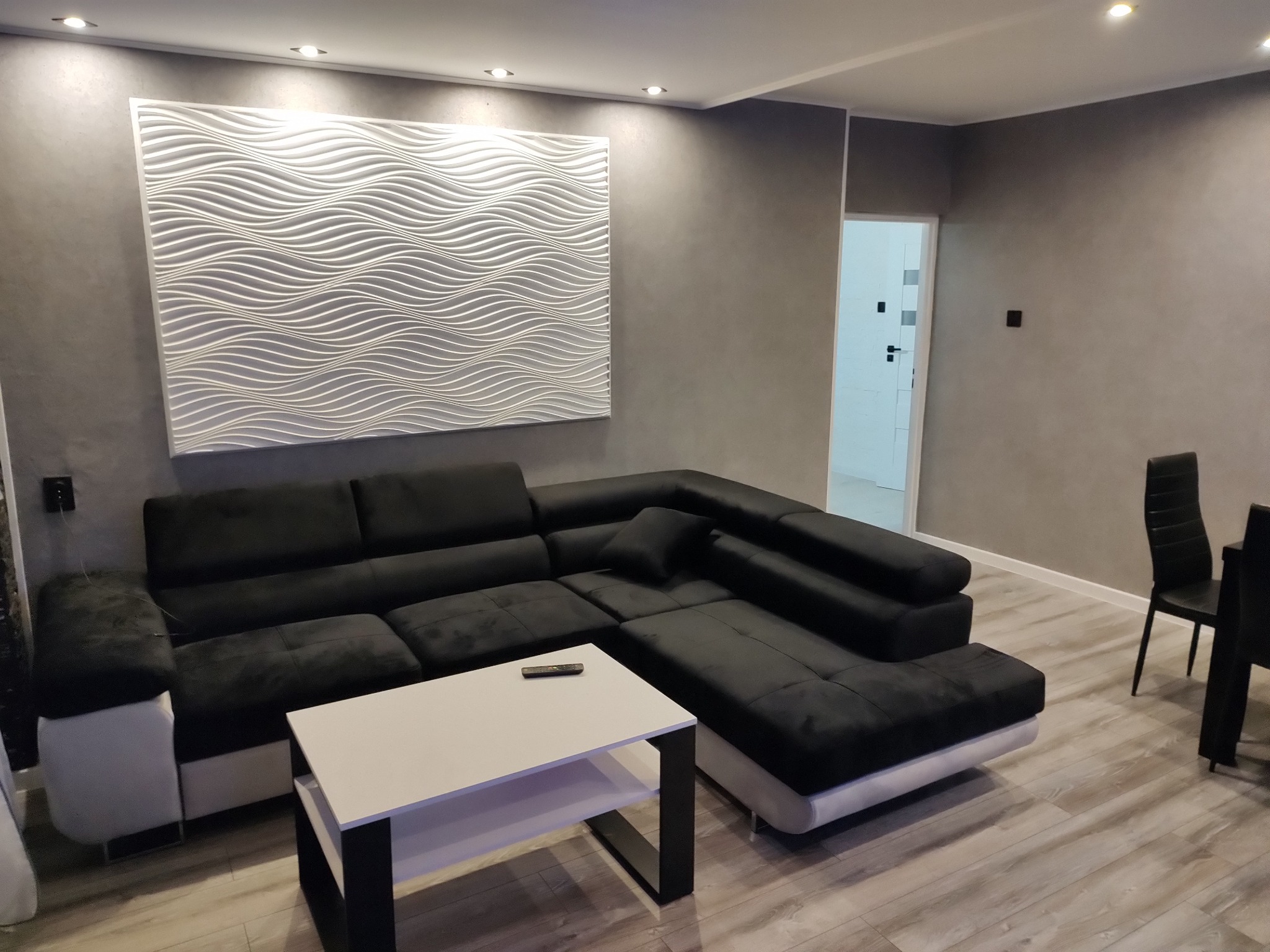 Szary salon z narożnikiem i dekoracyjną falistą płytą ścienną. Ściany wykończone w stylu betonu architektonicznego. Widoczne oświetlenie punktowe i minimalistyczny stolik.