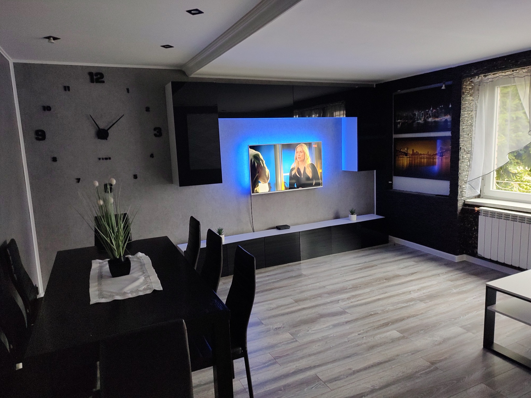 Nowoczesne wnętrze salonu z dużym zegarem na szarej ścianie, podświetlonym telewizorem i ciemnymi meblami. Parkiet w jasnych odcieniach drewna.