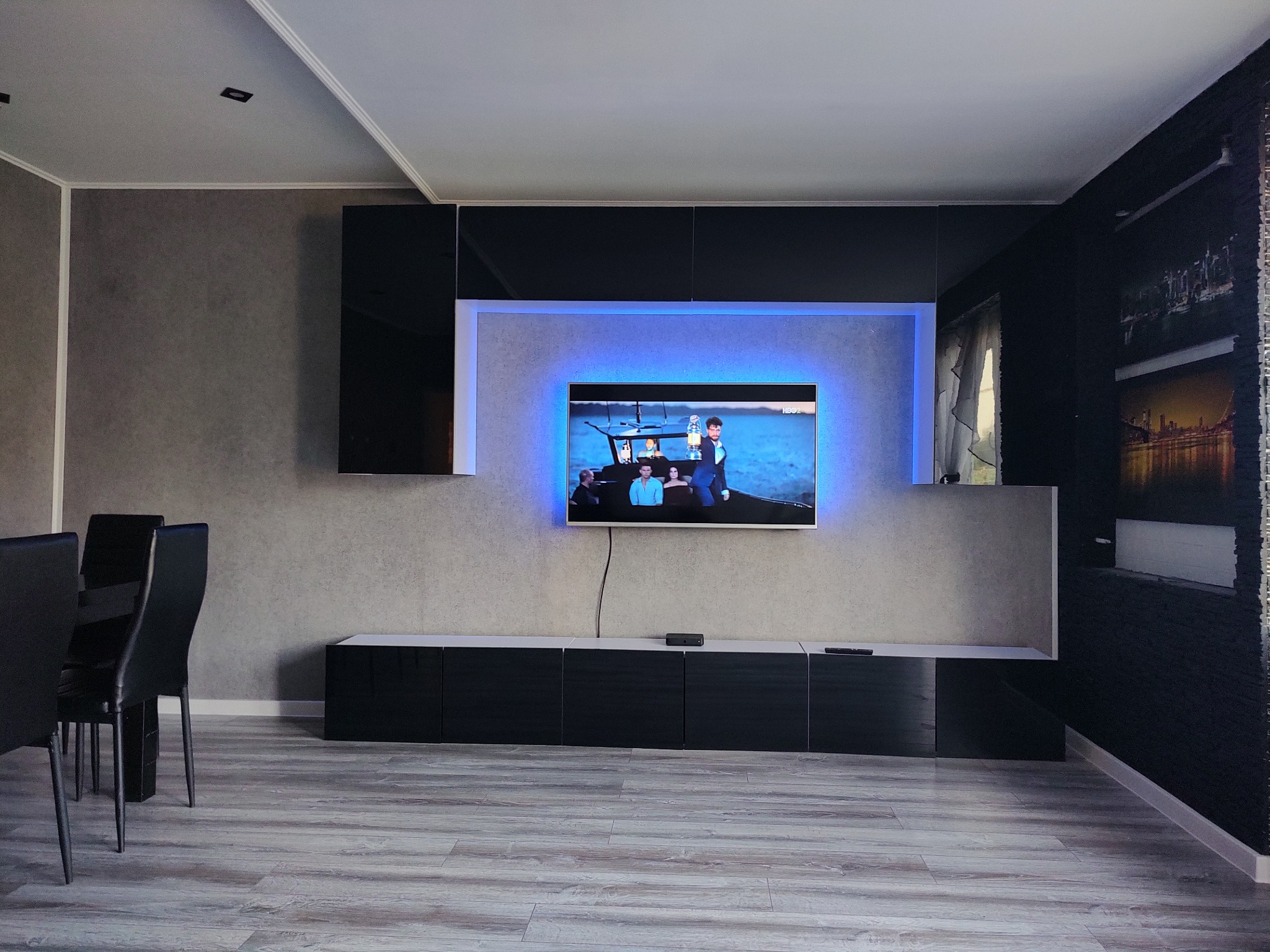 Nowoczesny salon z podświetlonym telewizorem, czarnymi meblami i szarą ścianą. Widoczne krzesła i panele podłogowe. Oświetlenie LED za telewizorem.