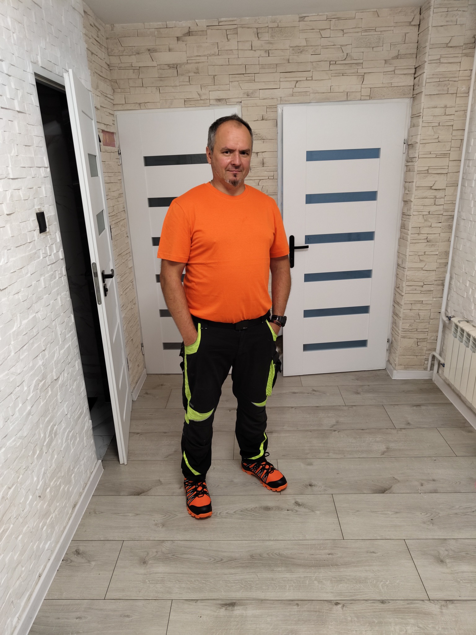 Mężczyzna w stroju roboczym stoi na tle wykończonego wnętrza z dwiema białymi, nowoczesnymi drzwiami i ścianą z dekoracyjnej cegły. Widoczna grzejnik i podłoga.