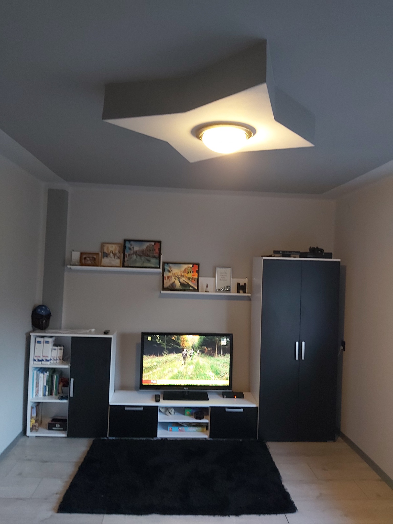 Jasne wnętrze pokoju z szarą ścianą, telewizorem i lampą w kształcie gwiazdy na suficie. Widoczne meble w odcieniach bieli i czerni oraz ciemny dywan.