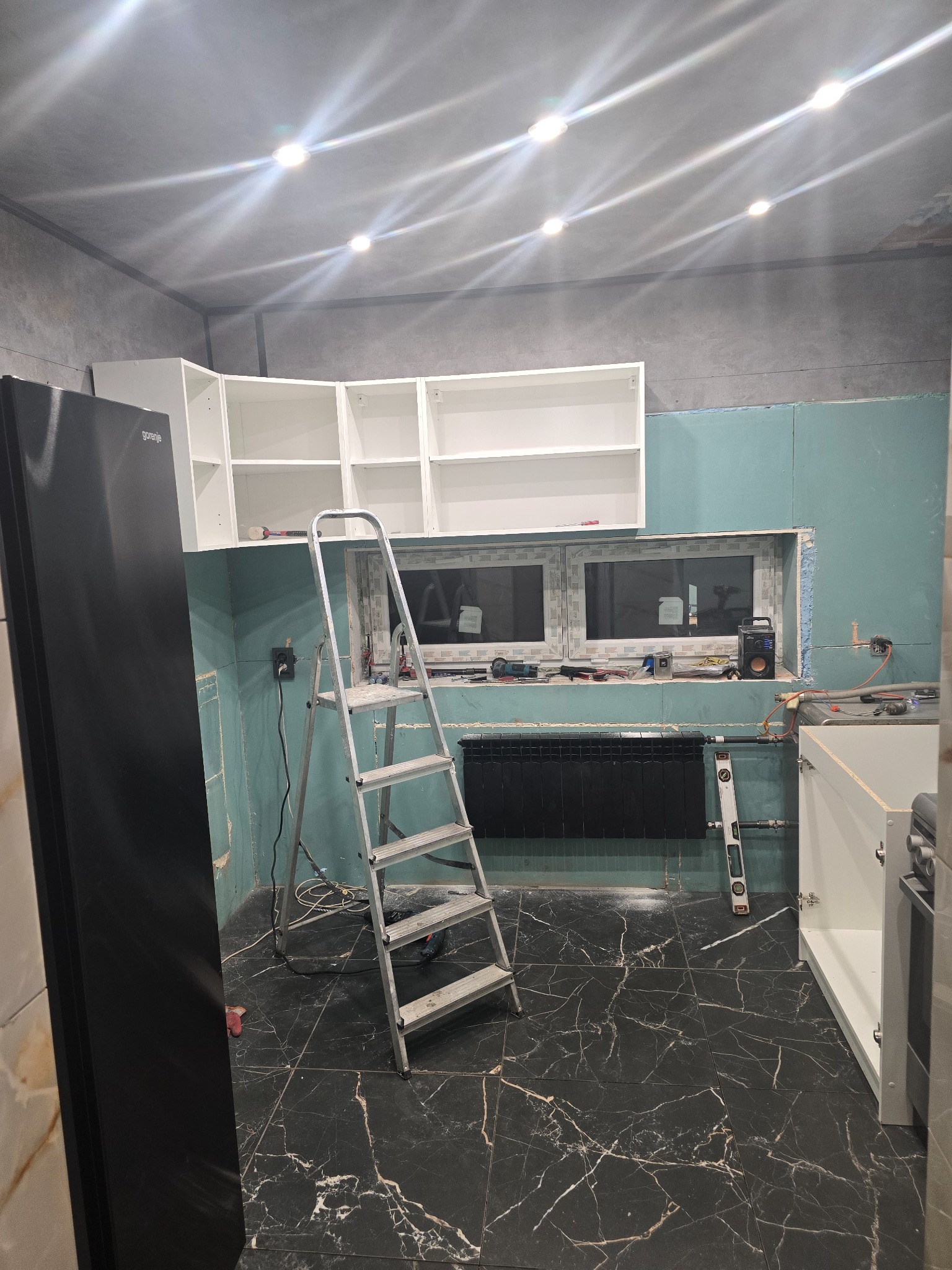 Kuchnia w trakcie remontu: zamontowane białe szafki wiszące, ściany z płyt gipsowo-kartonowych, drabina aluminiowa, czarna marmurowa podłoga i widoczne narzędzia.