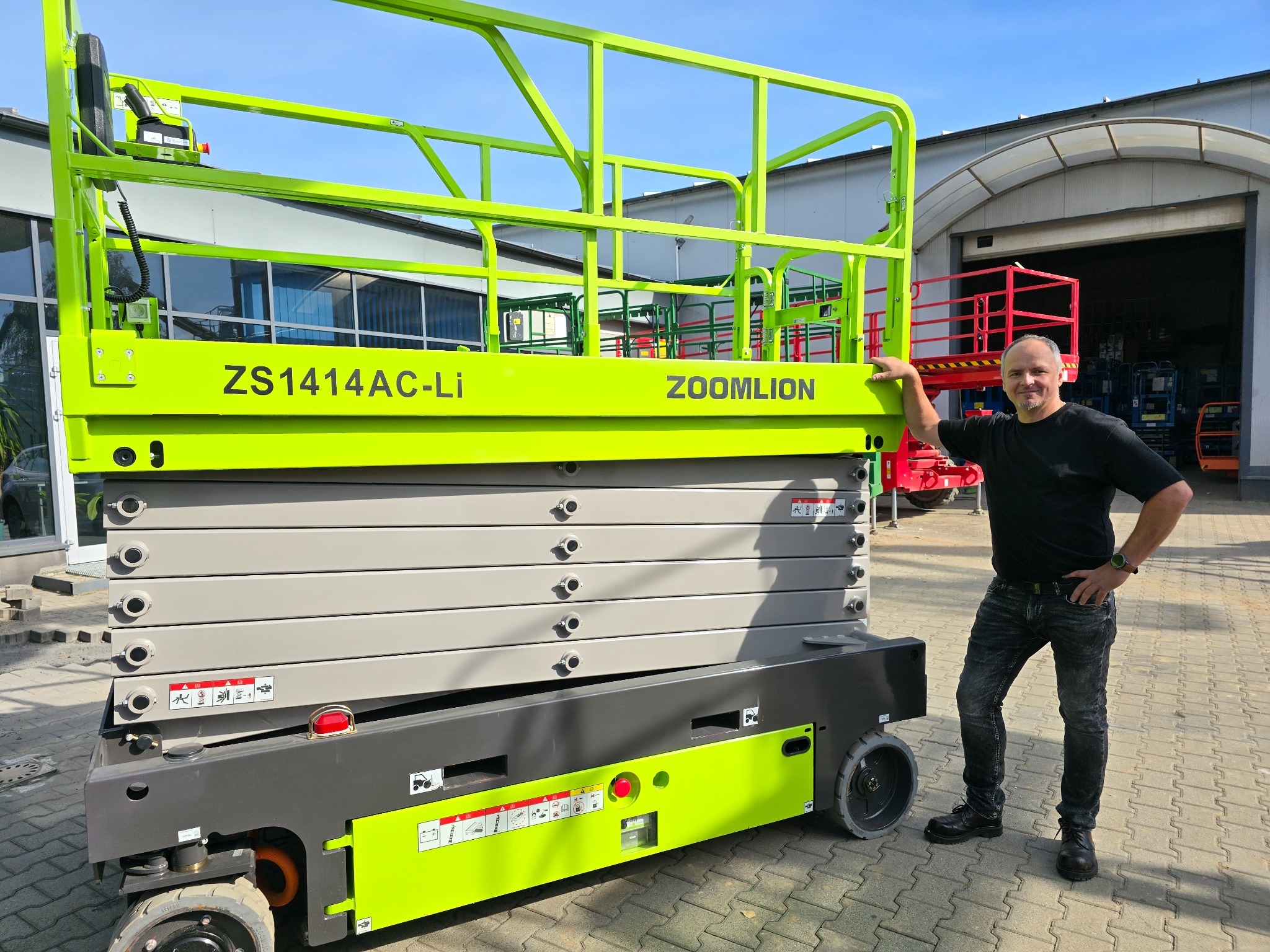 Mężczyzna opiera się o podnośnik nożycowy ZS1414AC-Li Zoomlion na zewnątrz, z widocznym fragmentem hali w tle i kostką brukową.