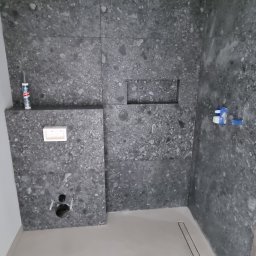 Dan-tech Daniel Gil - Szara łazienka w trakcie wykańczania: ułożone płytki imitujące kamień na ścianach, zamontowany odpływ liniowy, widoczne elementy instalacji. Surowy, nowoczesny styl.