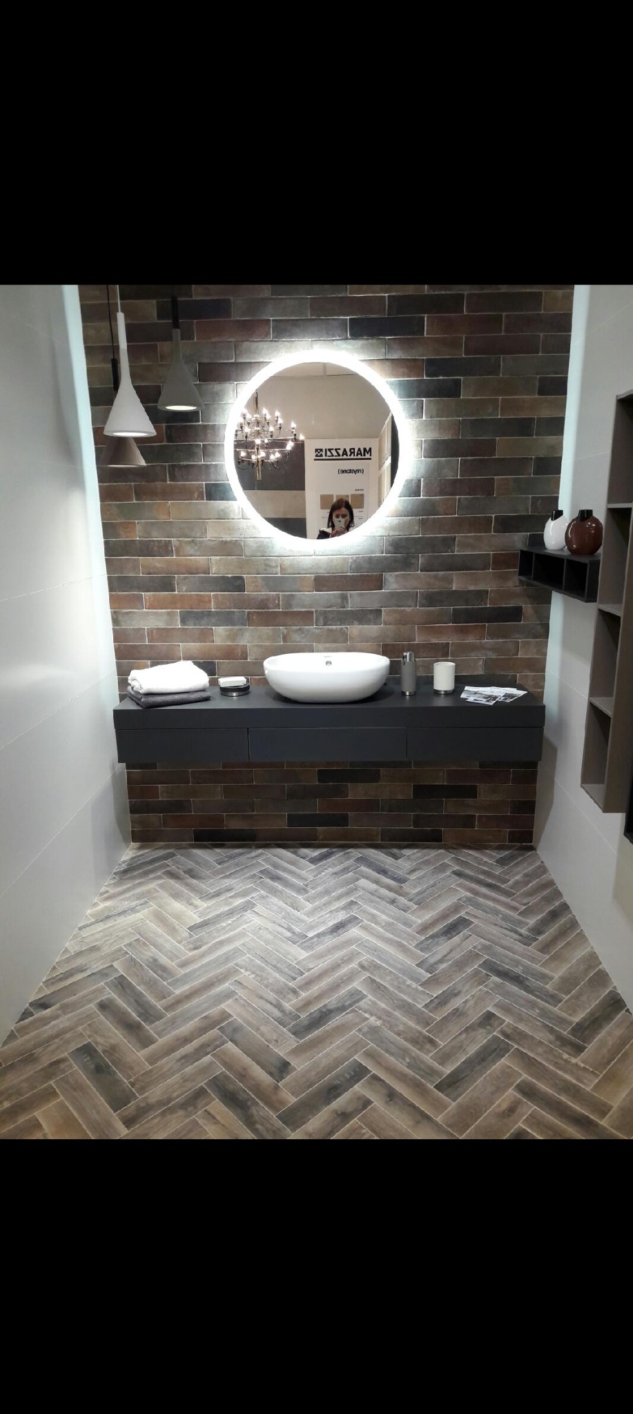 Stylowa łazienka z umywalką nablatową, okrągłym lustrem z podświetleniem LED i oryginalną, ceglaną ścianą. Na podłodze płytki ułożone w jodełkę.