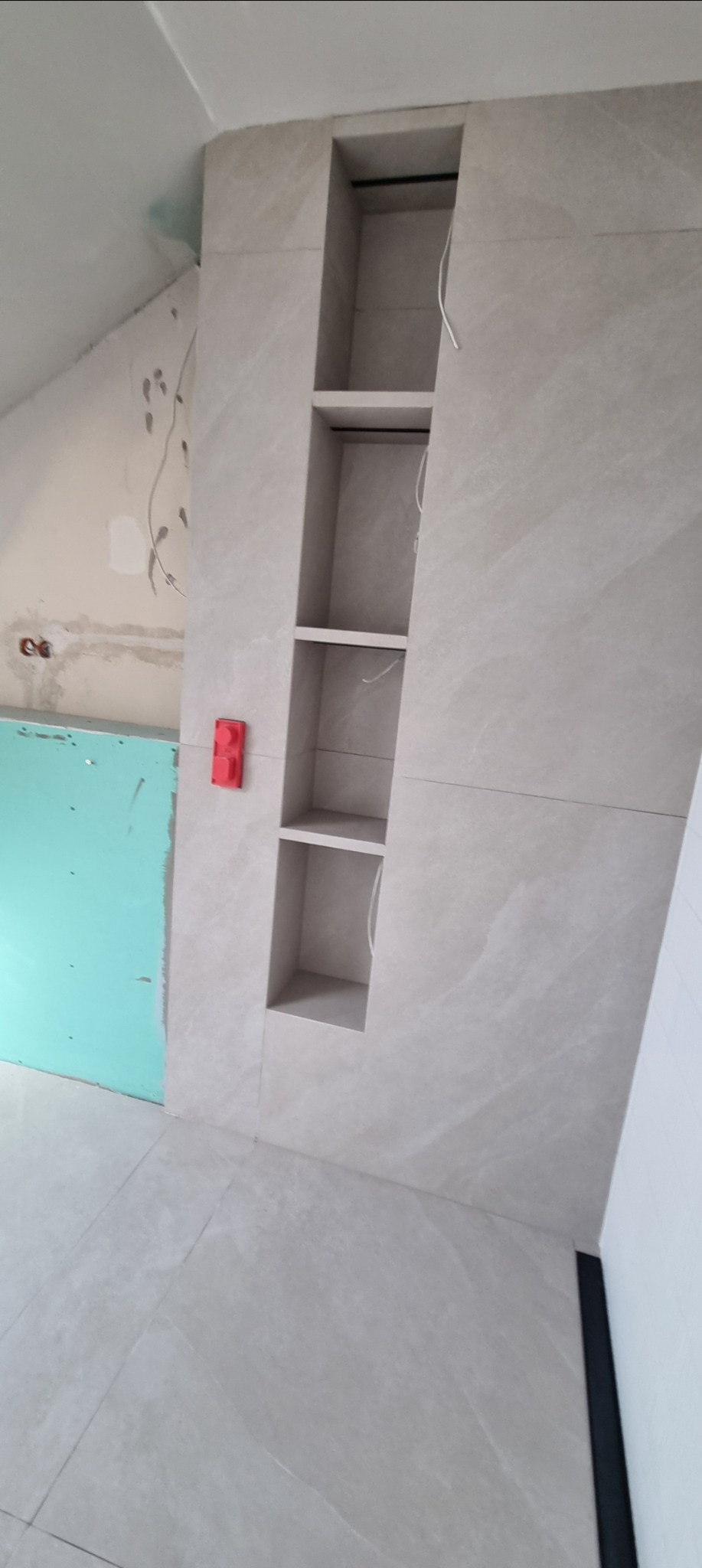Wykończona ściana z wnękami wyłożona płytkami imitującymi kamień, widoczne fragmenty instalacji elektrycznej i płyty gipsowej na ścianie. Szara podłoga z płytek.