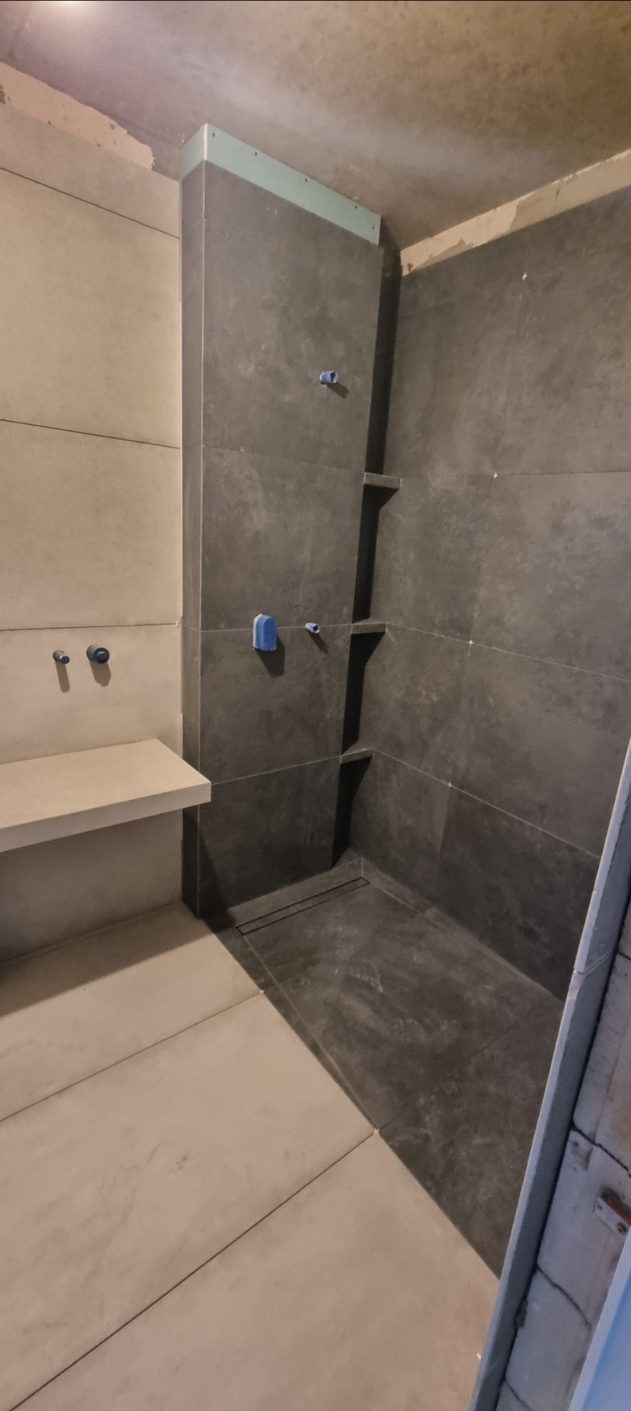 Nowoczesna łazienka w trakcie wykańczania: szare płytki, półki wnękowe w strefie prysznica, jasny blat umywalkowy. Widoczne niebieskie zaślepki na instalacjach.