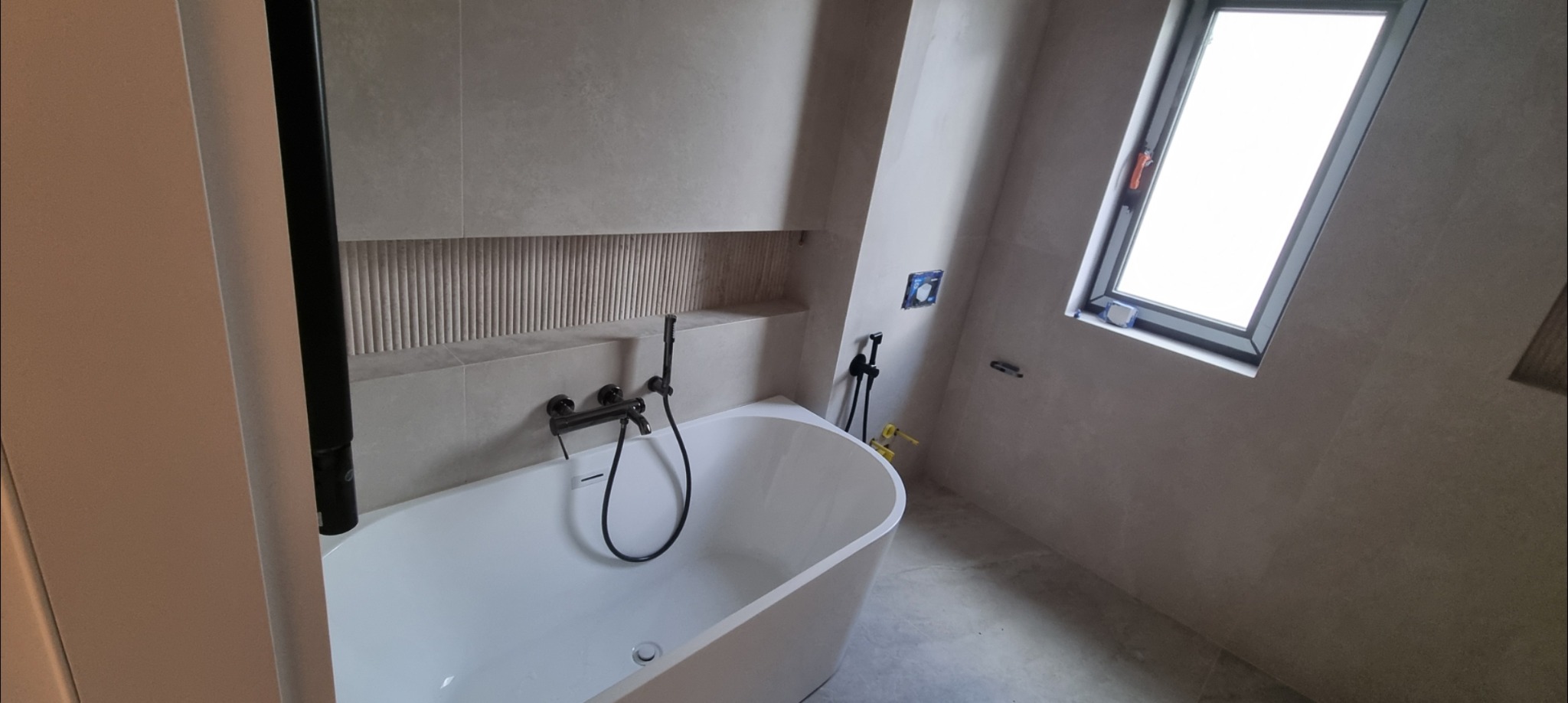 Nowoczesna łazienka w beżowych tonacjach: wolnostojąca wanna z czarną armaturą, dekoracyjna wnęka z ryflowanym wykończeniem, okno dachowe. Minimalistyczny design.