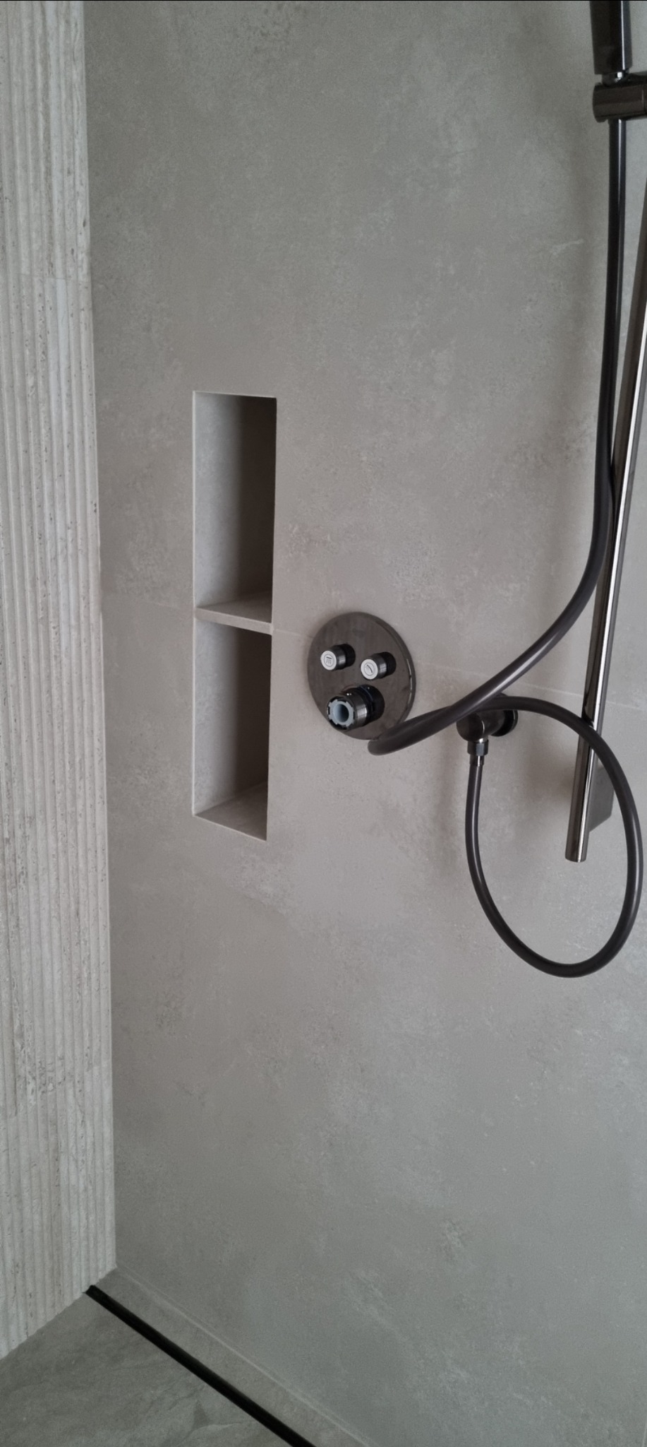 Minimalistyczna łazienka: szara płytka imitująca kamień, wnęka z półką, czarna armatura natryskowa, odpływ liniowy przy ścianie z dekoracyjną płytką.