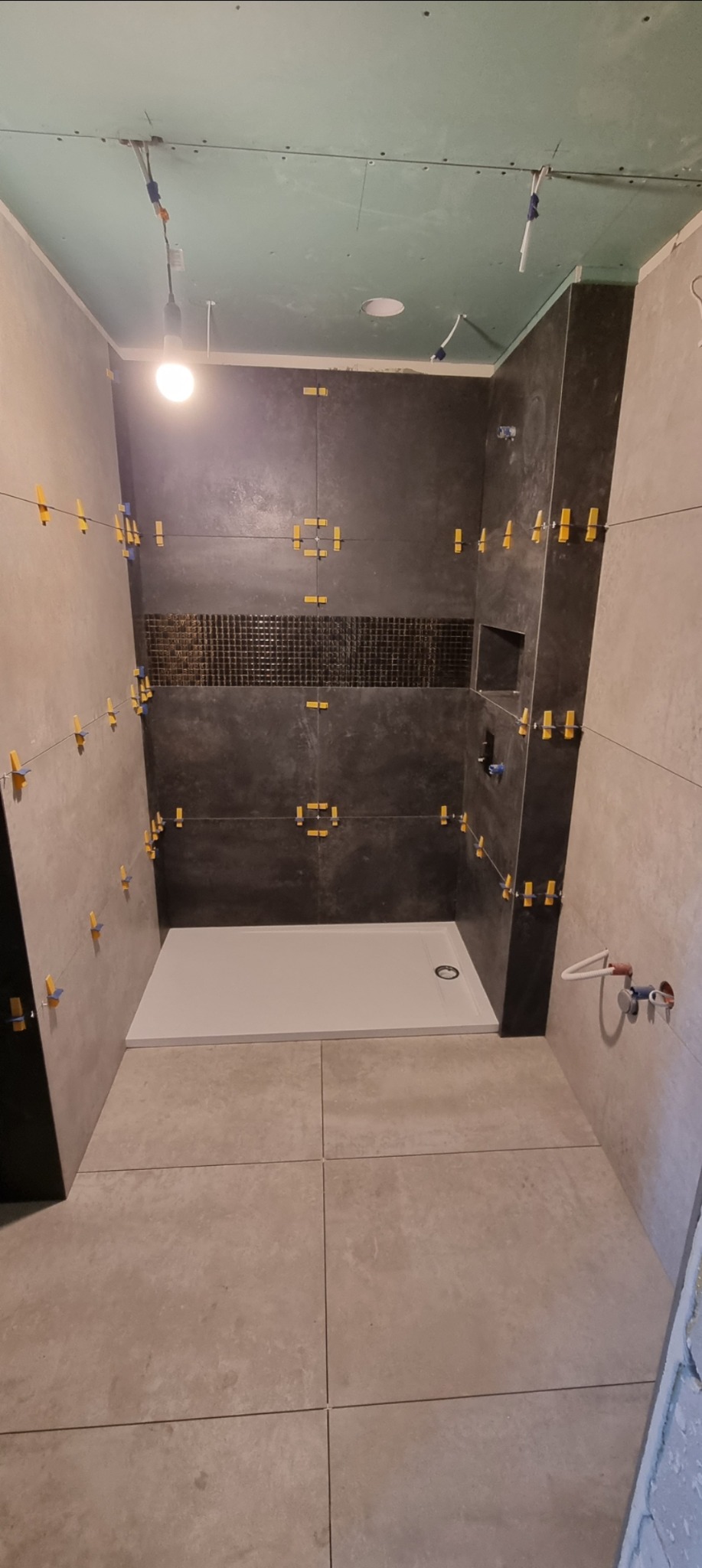 Łazienka w trakcie wykańczania: szare płytki na ścianach i podłodze, biały brodzik, żółte klipsy poziomujące płytki, surowy sufit.
