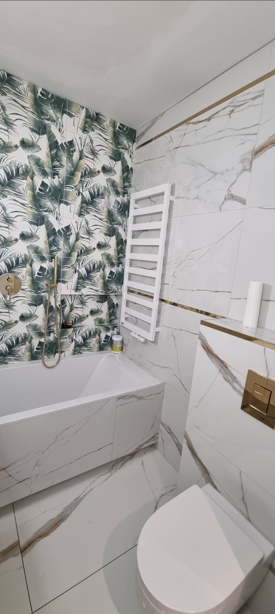 Wykończona łazienka z wanną, toaletą i grzejnikiem drabinkowym. Na ścianach białe płytki imitujące marmur z akcentami złota i tapeta w tropikalne liście.