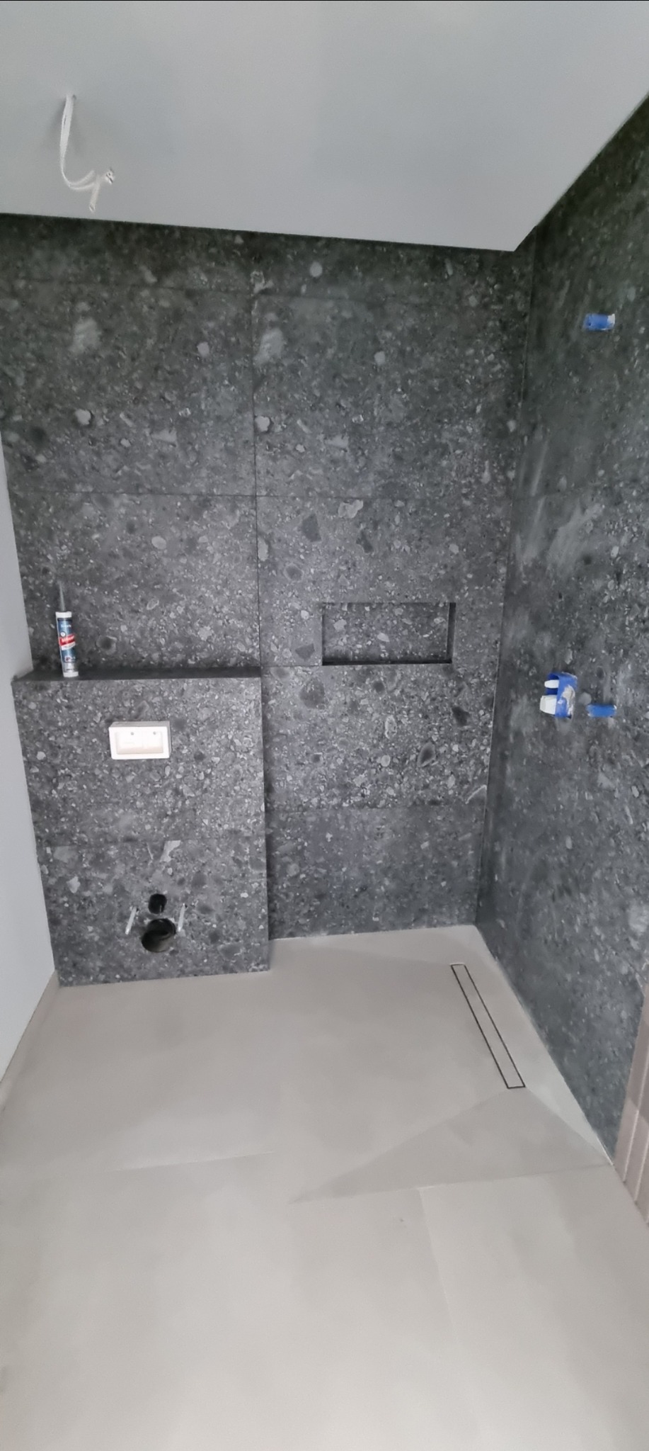 Szara łazienka w trakcie wykańczania: ułożone płytki imitujące kamień na ścianach, zamontowany odpływ liniowy, widoczne elementy instalacji. Surowy, nowoczesny styl.