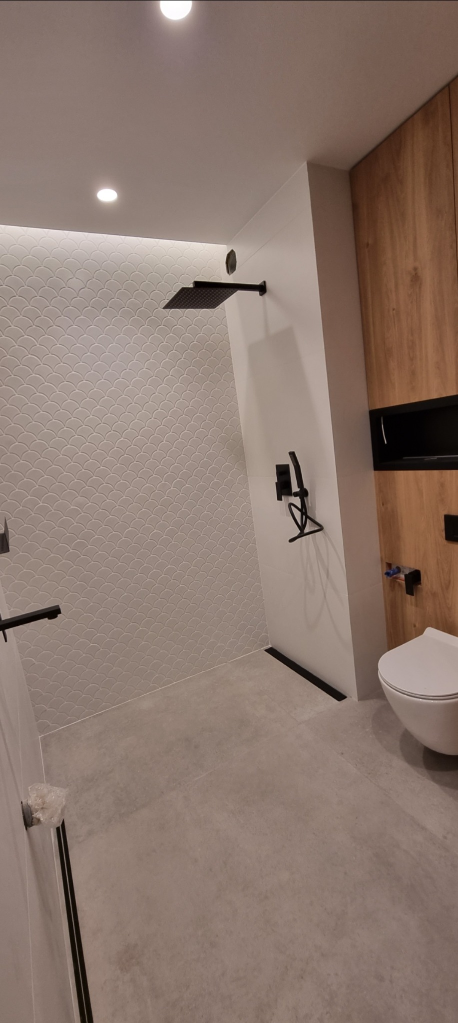Nowoczesna łazienka z kabiną typu walk-in, wykończona płytkami w kształcie rybiej łuski i czarną armaturą. Widoczna toaleta i drewniane szafki.