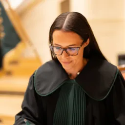 Portret kobiety adwokat w todze, piszącej dokument na drewnianym biurku. Ujęcie z bliska, skupione na detalu stroju i czynności pisania. Stonowana kolorystyka.