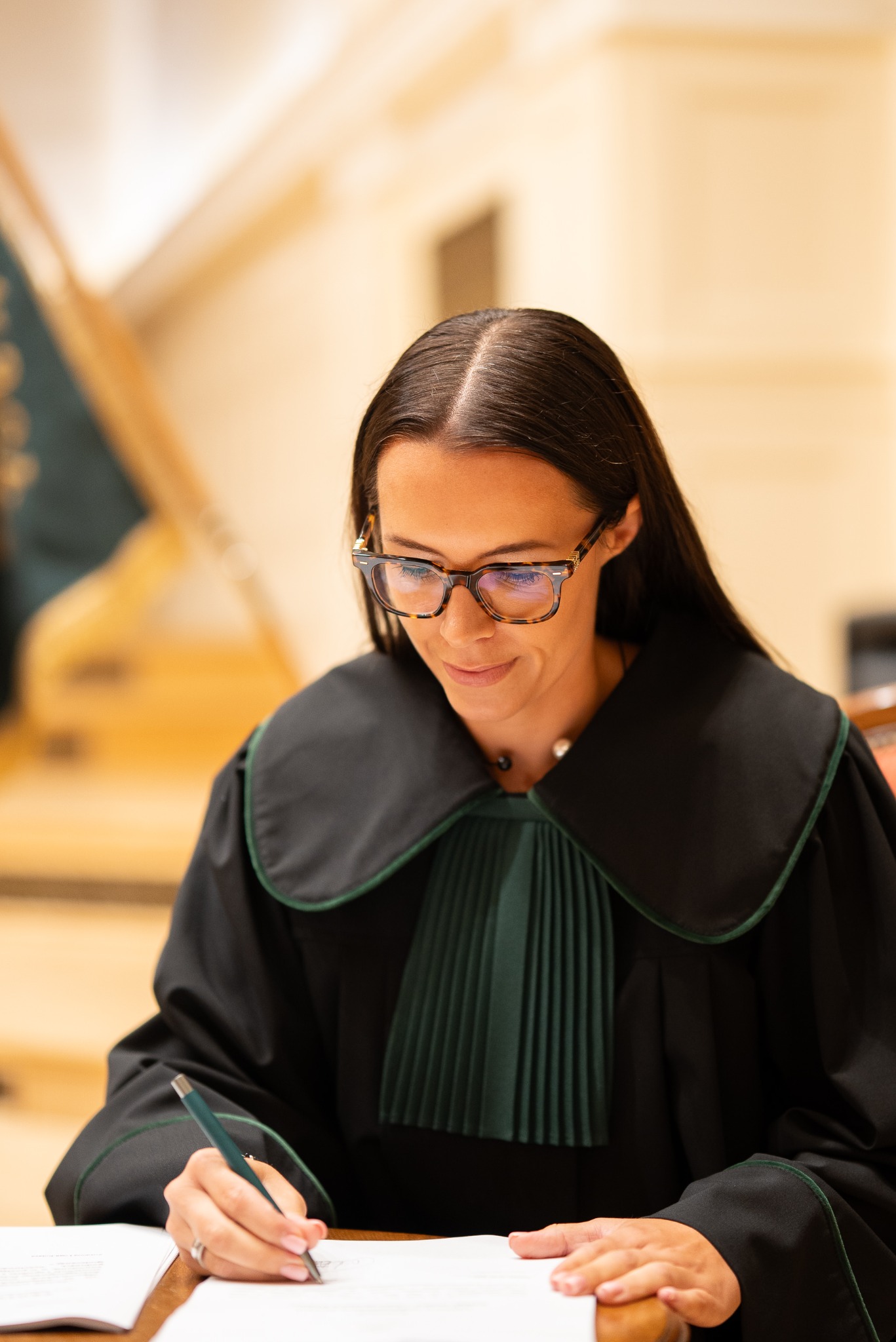 Portret kobiety adwokat w todze, piszącej dokument na drewnianym biurku. Ujęcie z bliska, skupione na detalu stroju i czynności pisania. Stonowana kolorystyka.