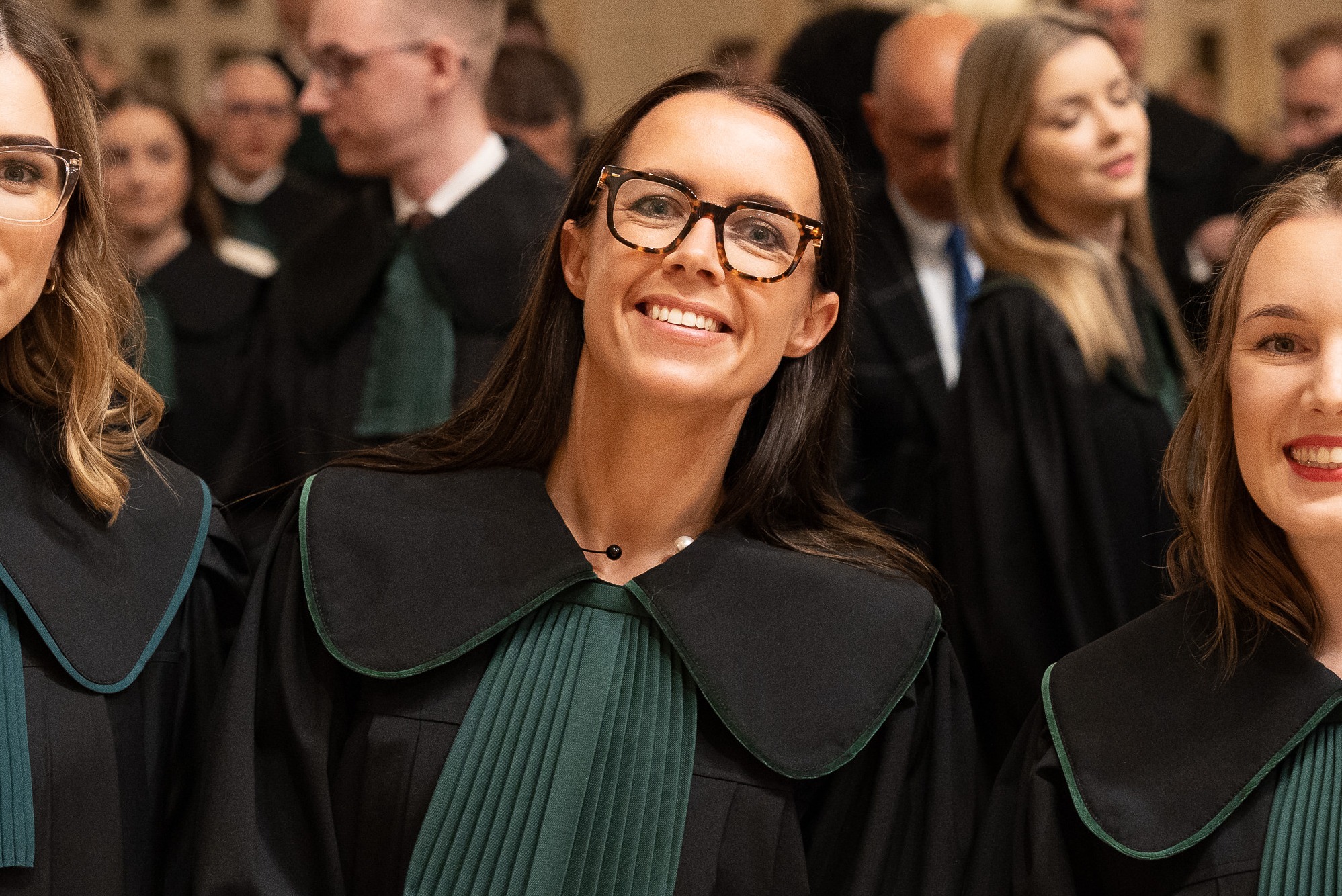 Uśmiechnięta kobieta w todze adwokackiej i okularach na tle grupy prawników podczas uroczystości ślubowania, widoczna z bliska.