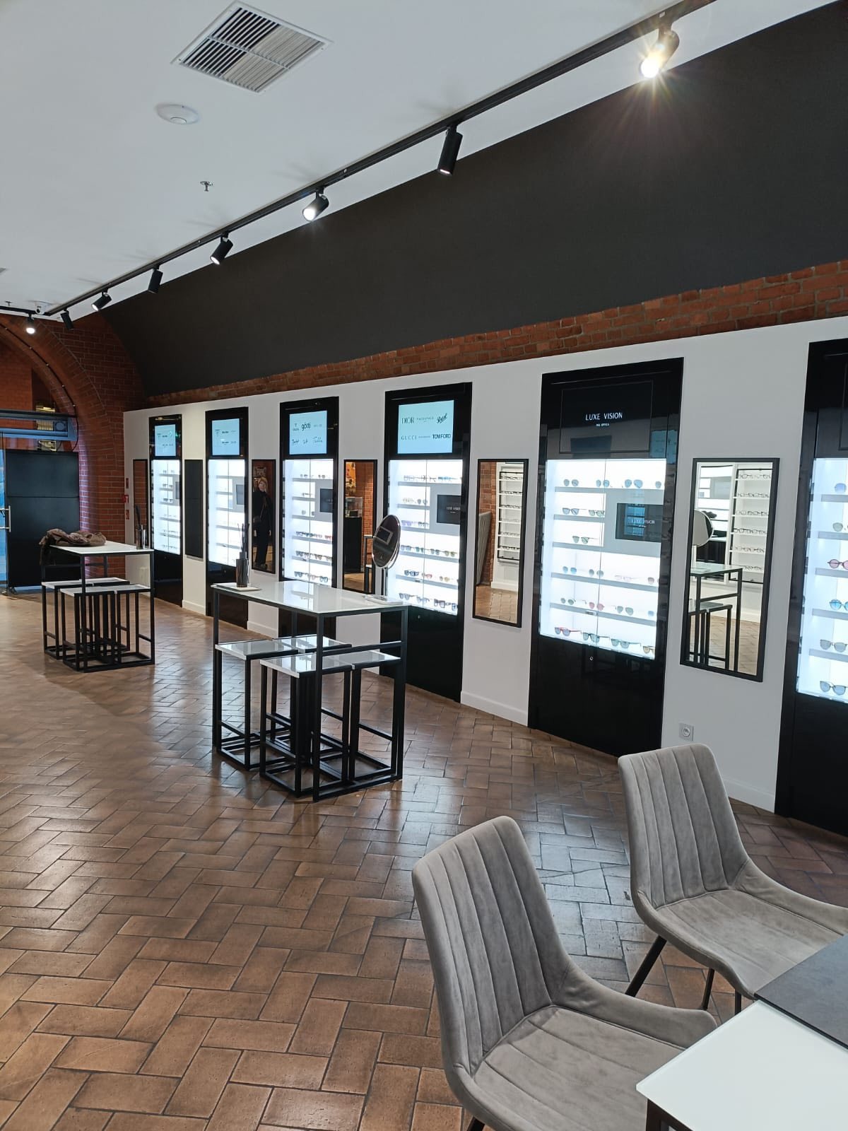 Wnętrze salonu optycznego z ekspozycją okularów w podświetlanych gablotach, stolikami i krzesłami. Ceglane akcenty na ścianach i parkiet ułożony w jodełkę.