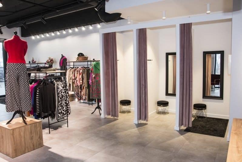 Jasne wnętrze butiku odzieżowego z trzema przymierzalniami z zasłonami w kolorze mauve. W tle wieszaki z ubraniami i manekin z czerwoną bluzką i wzorzystymi spodniami.