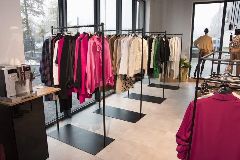 Rzędy czarnych, metalowych wieszaków na ubrania w butiku. Kolekcja odzieży w różnych kolorach. W tle widoczne okna i manekiny. Nowoczesny design wnętrza sklepu.