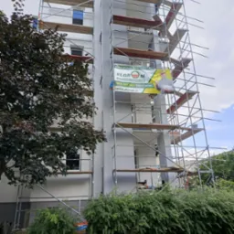 Ocieplony blok mieszkalny w rusztowaniach, z banerem reklamowym dostawcy materiałów budowlanych Elgrom, widok z poziomu trawnika i zieleni.