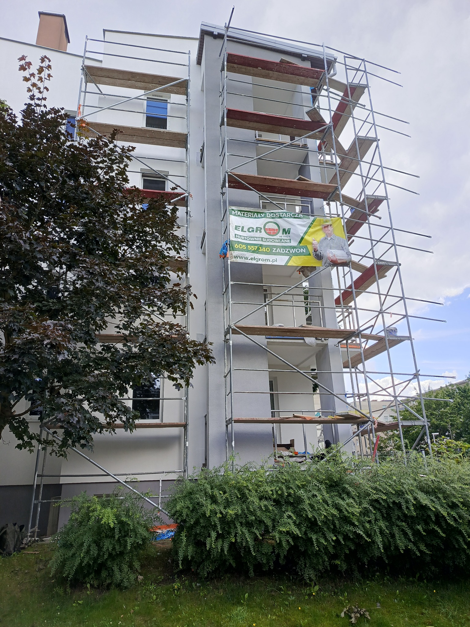Ocieplony blok mieszkalny w rusztowaniach, z banerem reklamowym dostawcy materiałów budowlanych Elgrom, widok z poziomu trawnika i zieleni.