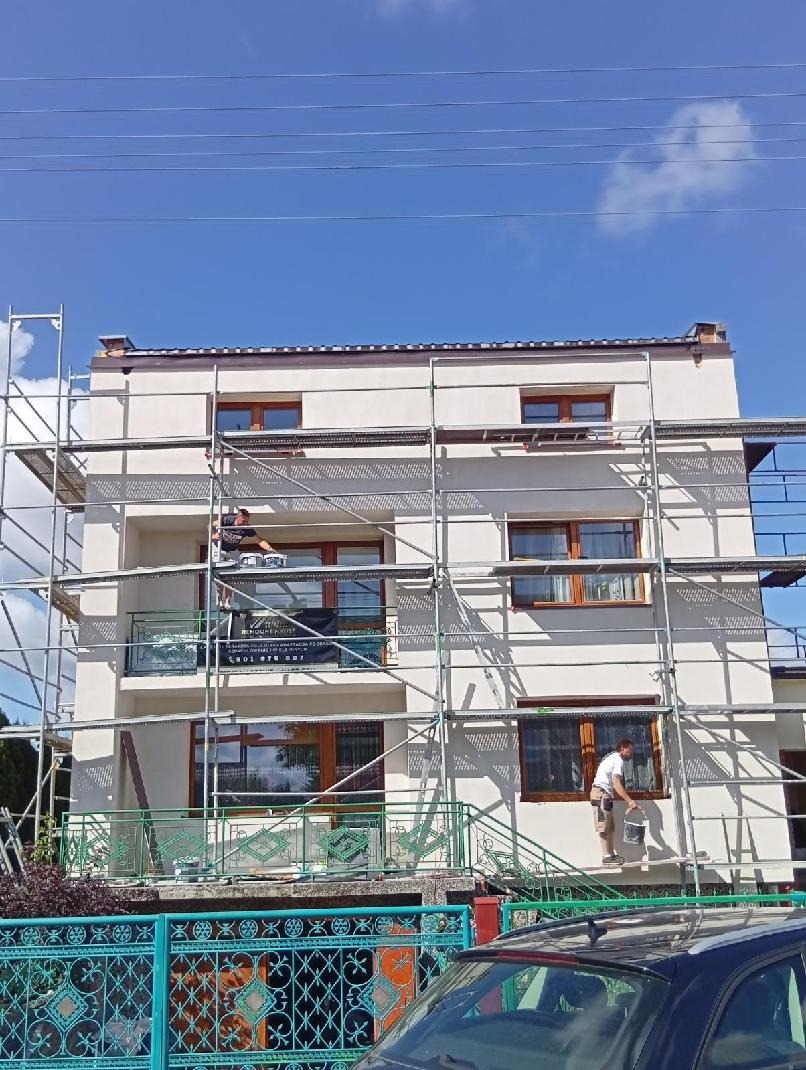 Dwóch pracowników na rusztowaniu maluje elewację trzypiętrowego domu. Widoczny fragment ogrodzenia i zaparkowany samochód przed budynkiem. Słoneczny dzień.
