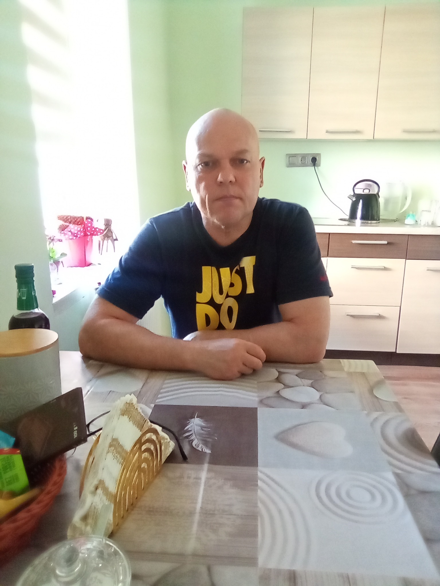 Mężczyzna siedzi przy stole w kuchni. W tle widać szafki kuchenne i okno. Na stole leżą różne przedmioty.