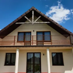 Dom z balkonem i drewnianą balustradą w Rejowcu Fabrycznym. Brązowe ramy okien, białe kolumny i beżowa elewacja. Widok na błękitne niebo z chmurami.