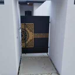 Rafał Adamczuk ALLStal - Nowoczesna, czarna furtka wejściowa z ozdobnymi, ażurowymi elementami w złotym kolorze, osadzona między białymi ścianami. Minimalistyczny design z geometrycznymi i roślinnymi motywami.