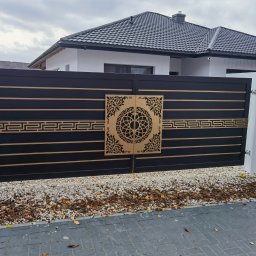 Rafał Adamczuk ALLStal - Nowoczesna, czarna brama wjazdowa z ażurowymi, złotymi zdobieniami, osadzona na białych słupkach. Widoczne kamienie i kostka brukowa na podjeździe.