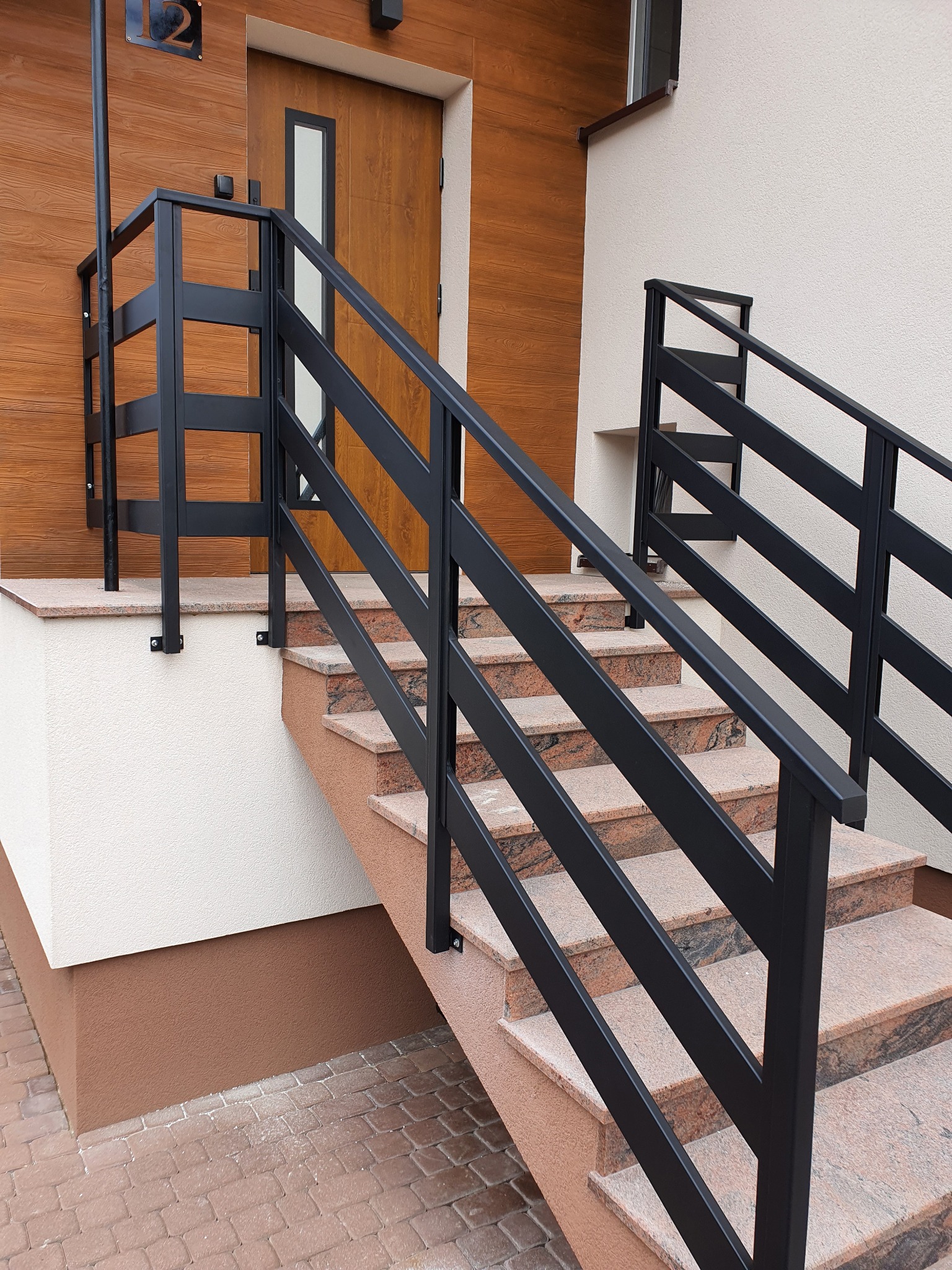 Nowoczesne, czarne balustrady metalowe przy schodach wejściowych do domu z drewnianą elewacją. Minimalistyczny design, proste linie. Kamienne stopnie schodów.