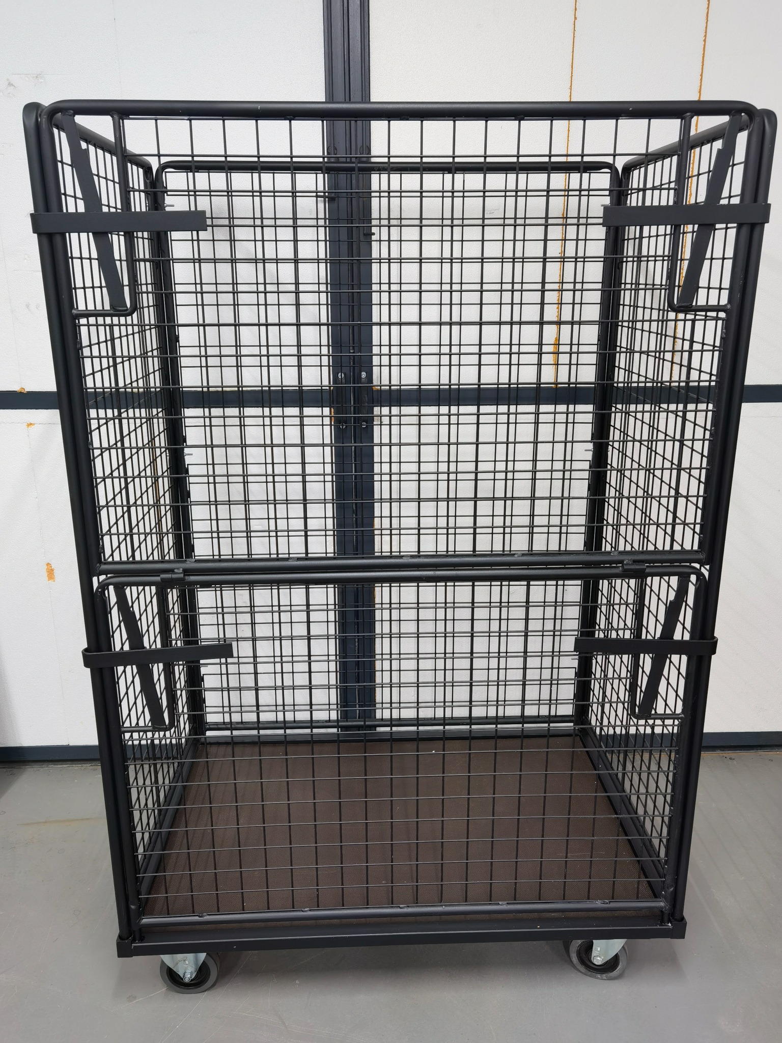 Czarny metalowy wózek z siatki na kółkach, z uchwytami z boku, stoi na szarej podłodze na tle białej ściany z ciemnymi panelami.