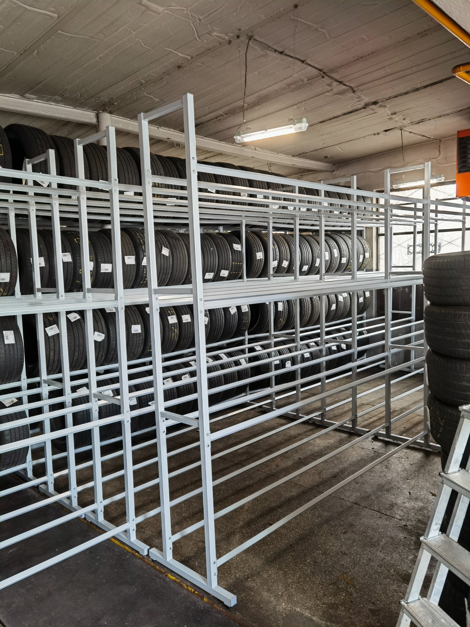 Metalowa konstrukcja regału na opony w warsztacie. Widoczne opony z etykietami, drabina aluminiowa. Szara podłoga i sufit z oświetleniem.