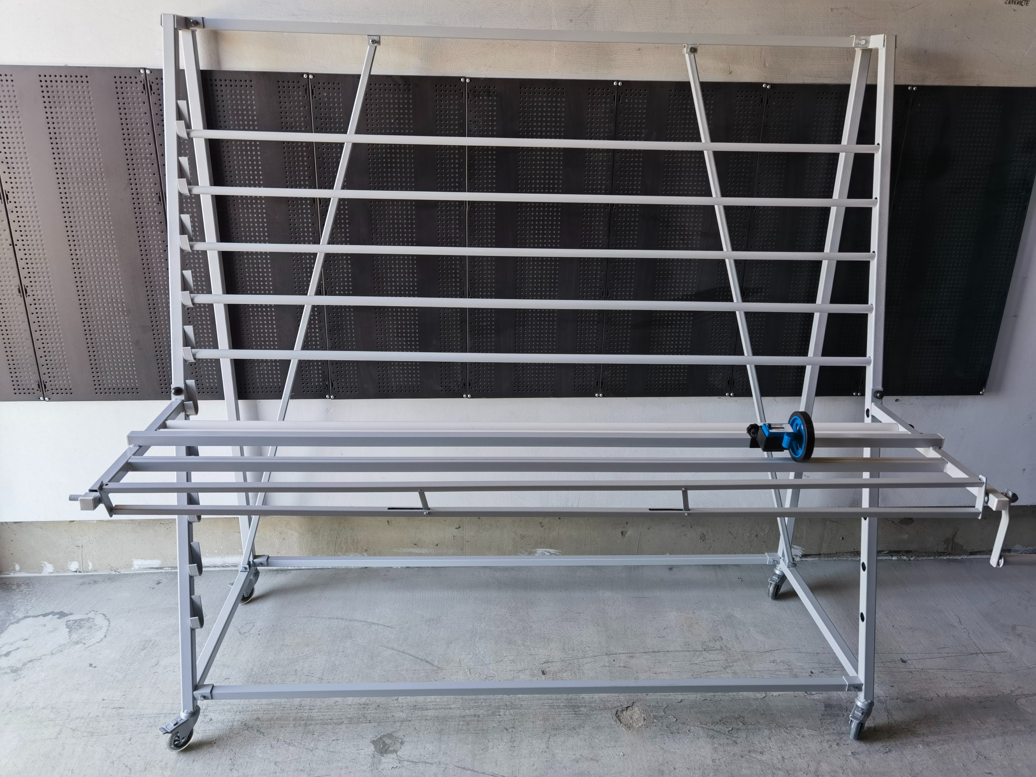 Stół warsztatowy z metalową konstrukcją na kółkach i narzędziem do cięcia płytek, na tle ściany z perforowanymi panelami.