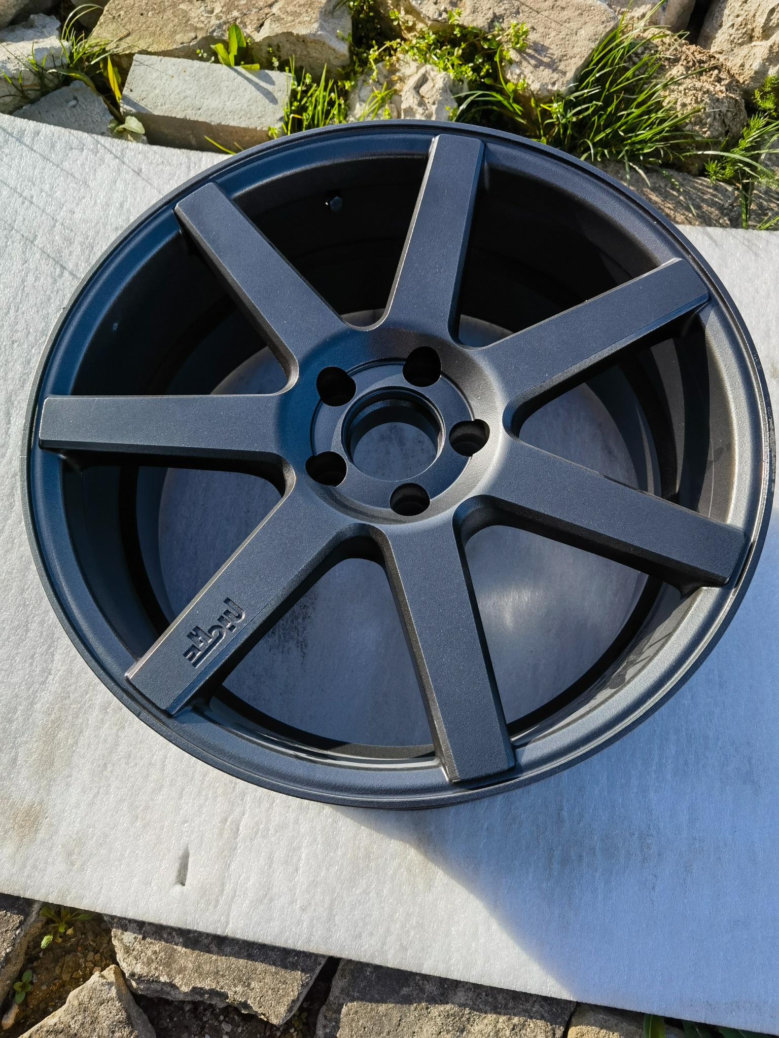 Szara felga aluminiowa z siedmioma ramionami na białym tle, z widocznym logo marki. Ujęcie z góry, z elementami otoczenia: kamienie i trawa.