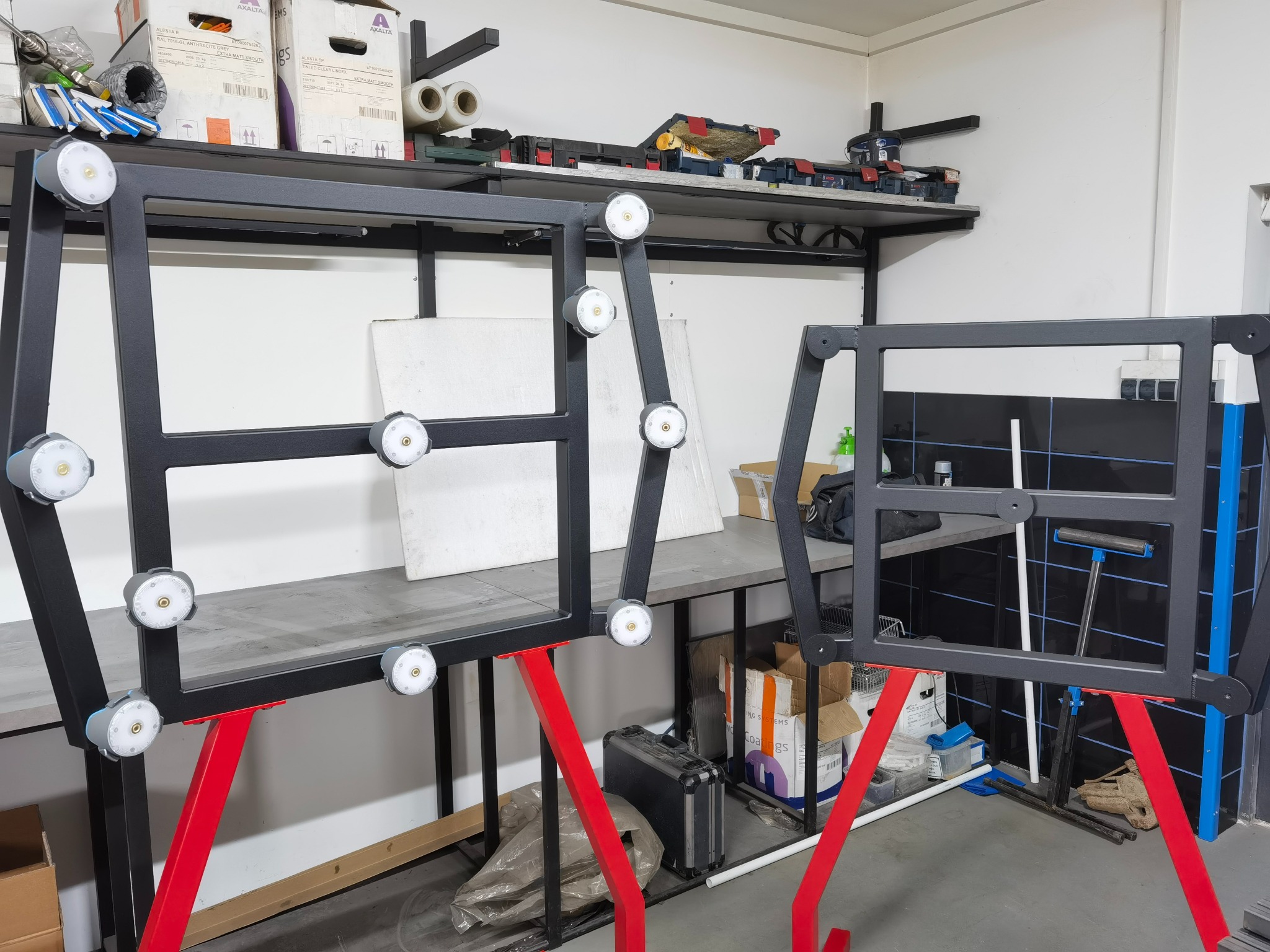 Dwie metalowe ramy z oświetleniem LED, pomalowane na czarno, wsparte na czerwonych nogach. W tle widoczne narzędzia i materiały warsztatowe.
