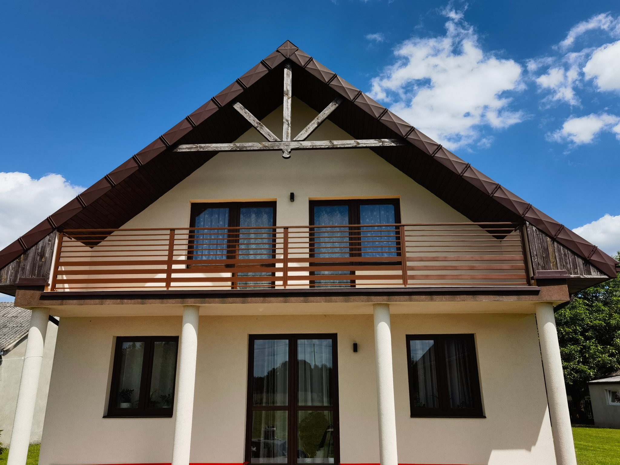 Dom z balkonem i drewnianą balustradą w Rejowcu Fabrycznym. Brązowe ramy okien, białe kolumny i beżowa elewacja. Widok na błękitne niebo z chmurami.