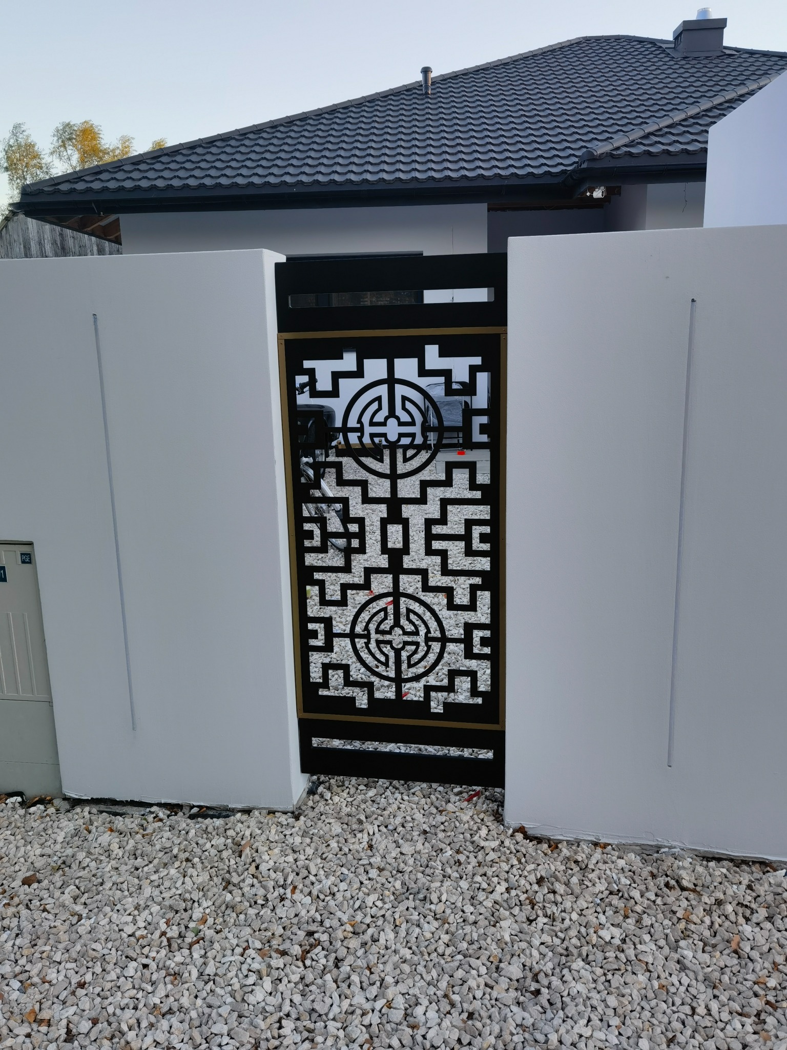 Nowoczesna brama wjazdowa z dekoracyjnym, geometrycznym wzorem w kolorze czarnym, osadzona między białymi, minimalistycznymi ścianami na tle szarej dachówki.