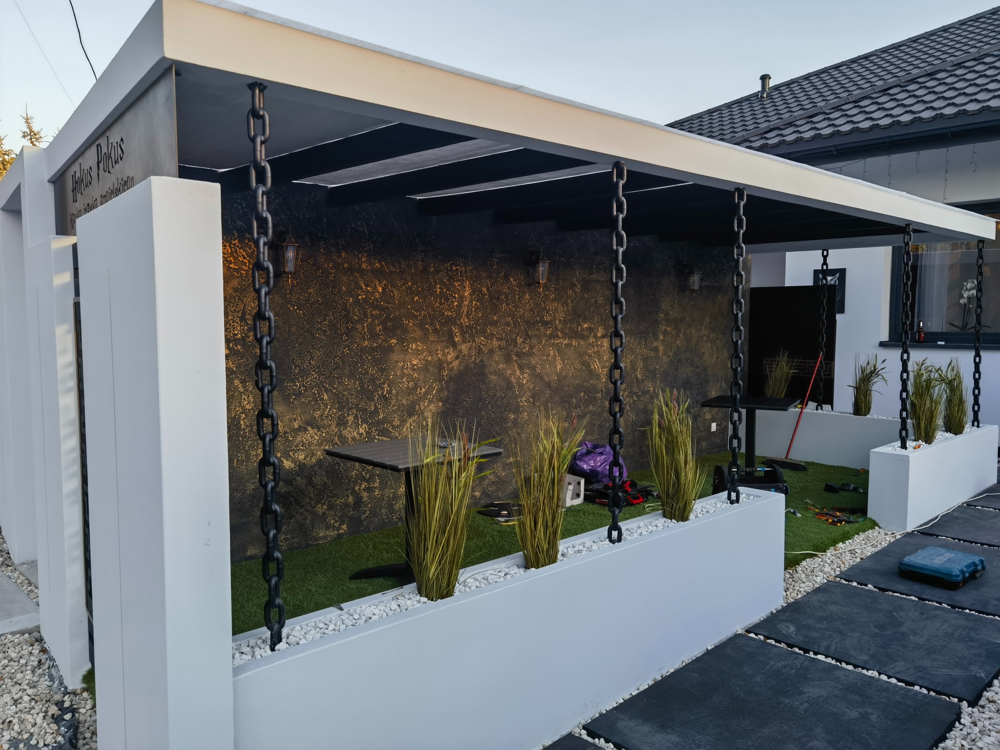 Nowoczesna pergola z metalowymi łańcuchami i dekoracyjną ścianą. Elementy małej architektury ogrodowej w kolorach bieli i czerni.