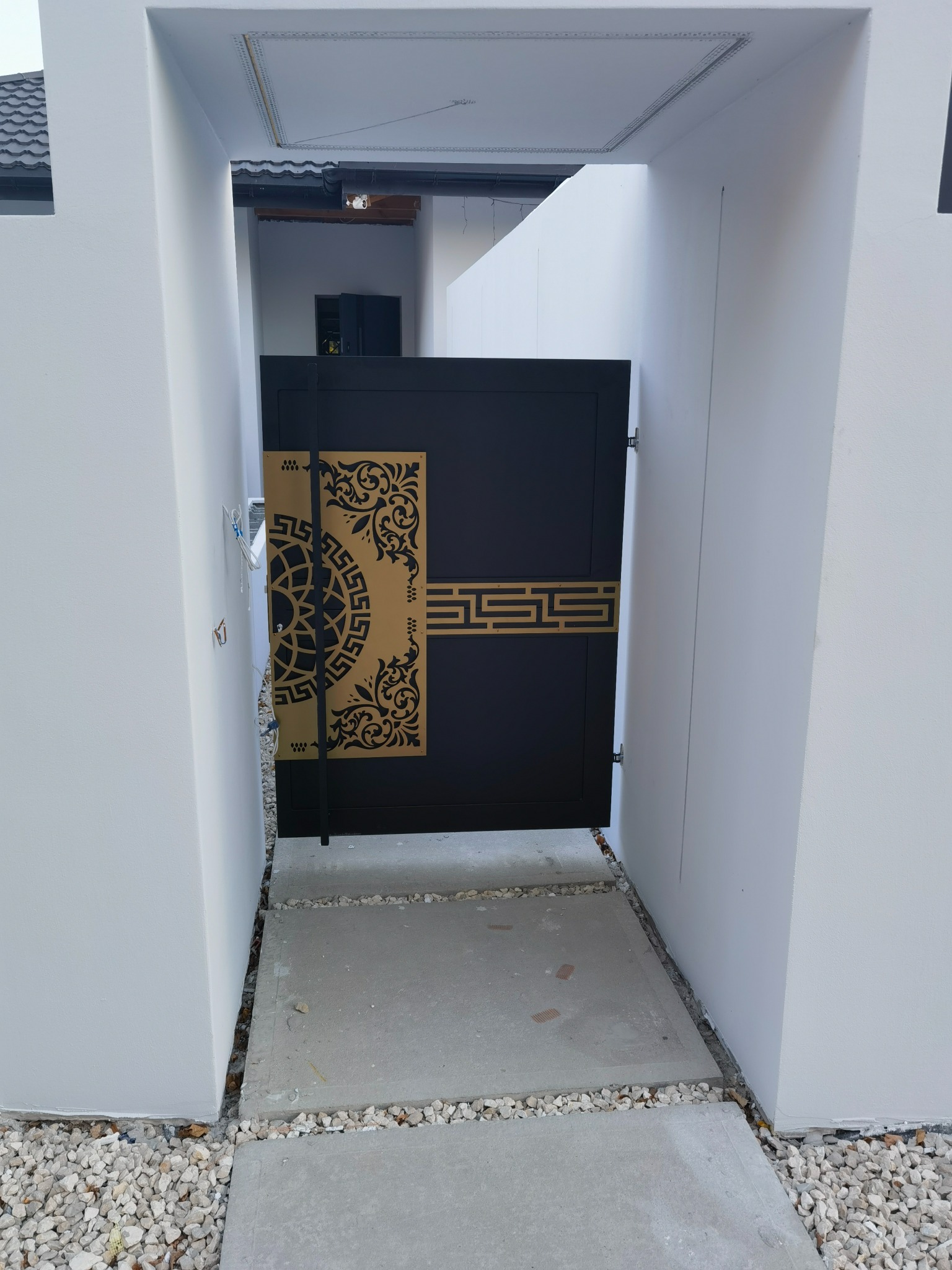 Nowoczesna, czarna furtka wejściowa z ozdobnymi, ażurowymi elementami w złotym kolorze, osadzona między białymi ścianami. Minimalistyczny design z geometrycznymi i roślinnymi motywami.