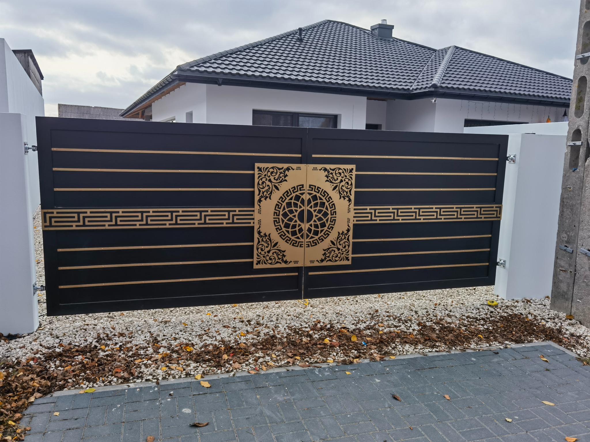 Nowoczesna, czarna brama wjazdowa z ażurowymi, złotymi zdobieniami, osadzona na białych słupkach. Widoczne kamienie i kostka brukowa na podjeździe.