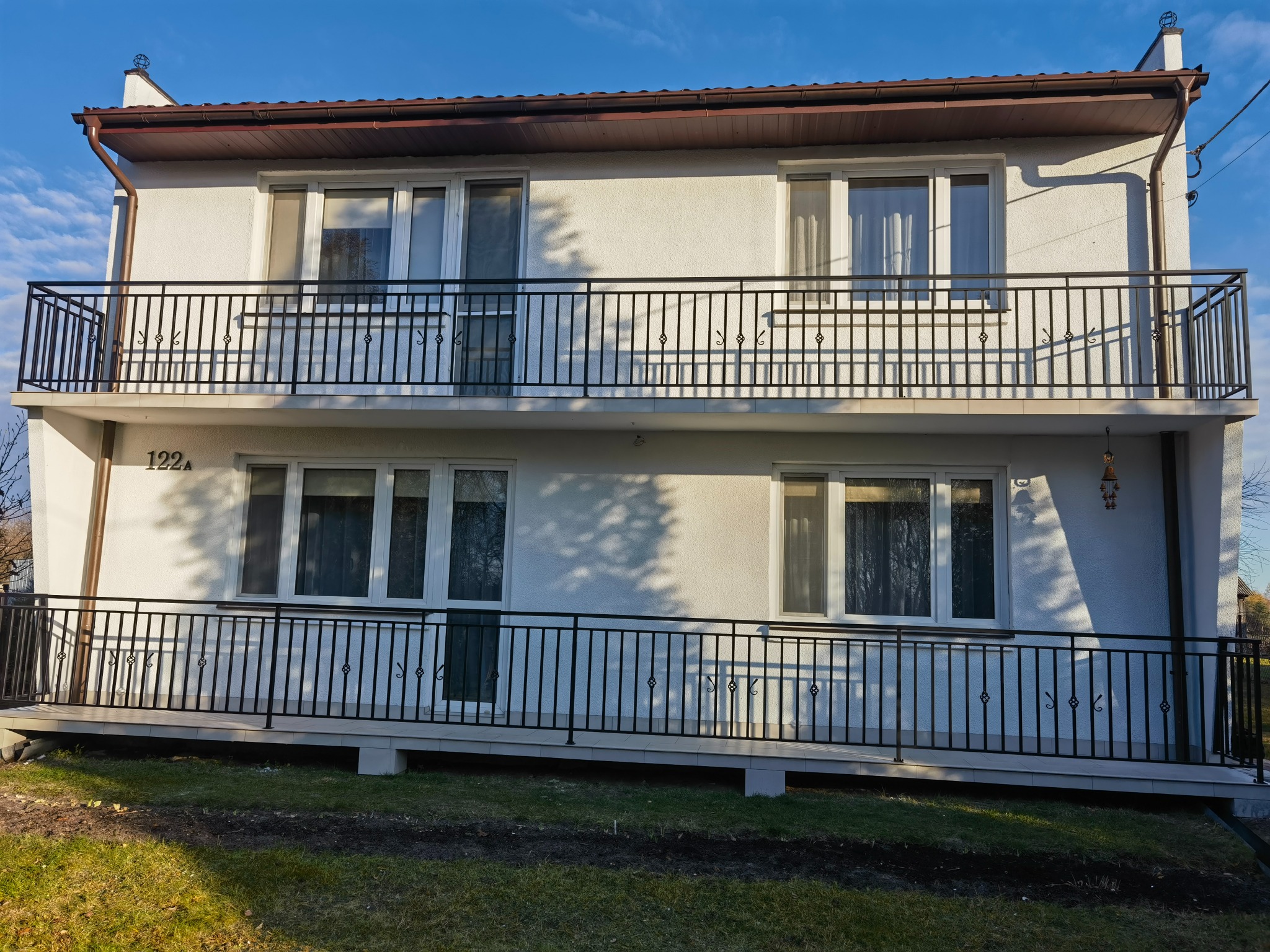 Dwukondygnacyjny budynek z czarnymi, metalowymi balustradami balkonowymi o dekoracyjnym wzorze, widziany w słoneczny dzień z perspektywy trawnika.