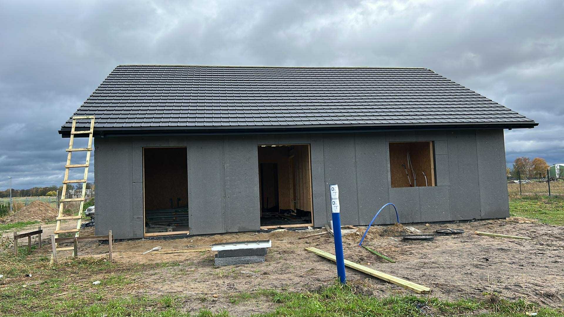 Dom w trakcie budowy z ciemnym dachem z dachówki, drewniana drabina oparta o ścianę, widoczne otwory okienne i drzwiowe, pochmurne niebo w tle.