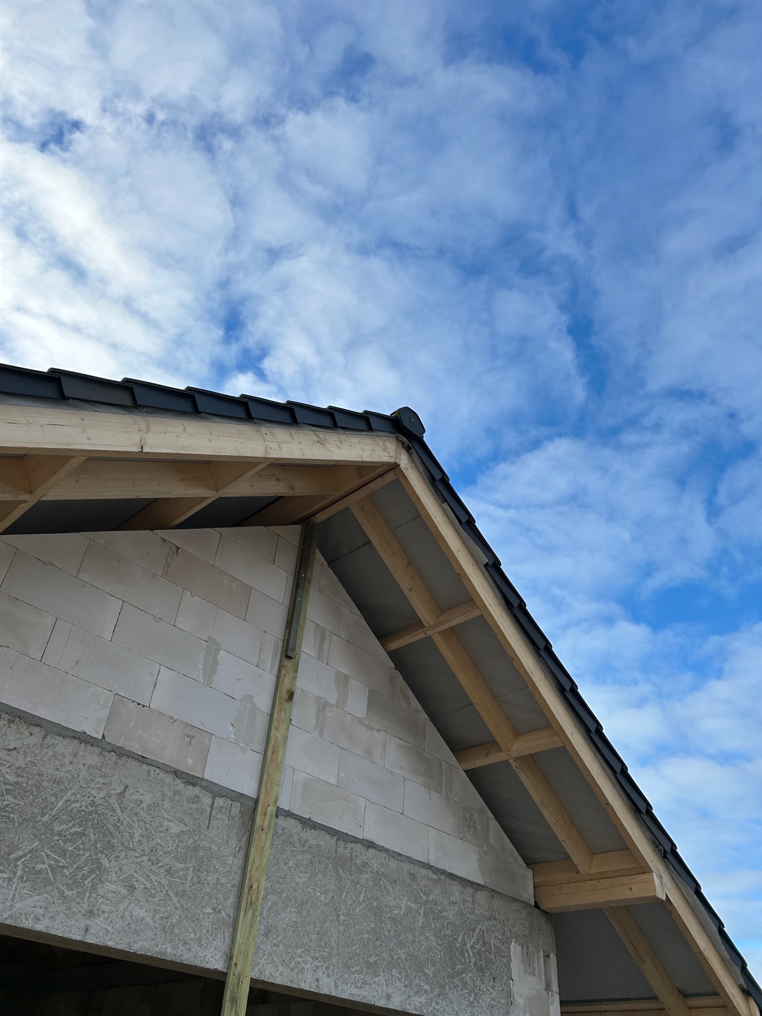 Fragment konstrukcji dachu z czarną dachówką na tle błękitnego nieba z chmurami. Widoczna drewniana więźba i fragment ściany z betonu komórkowego.