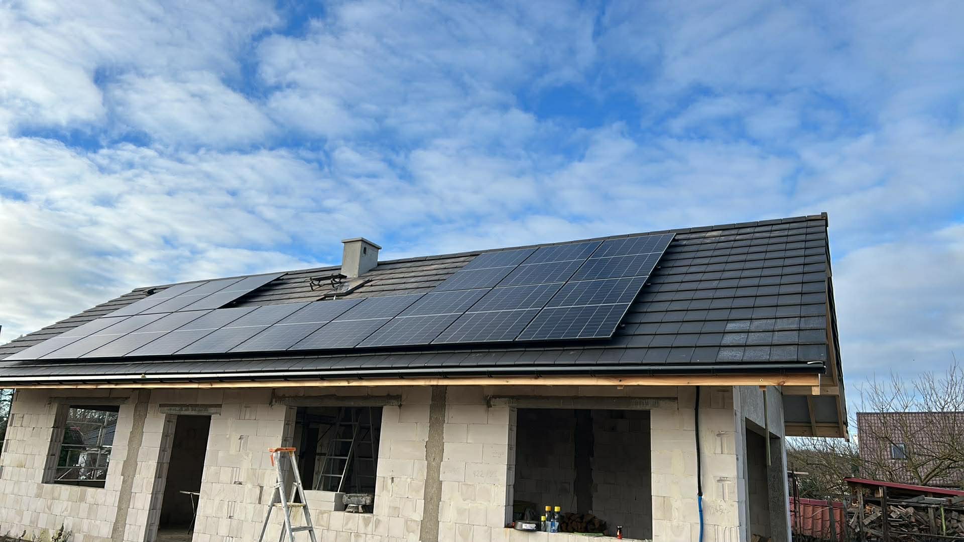 Dom w trakcie budowy z panelami słonecznymi na dachu z czarnej dachówki. Widoczne białe ściany z pustaków i drabina oparta o ścianę. Błękitne niebo z chmurami w tle.