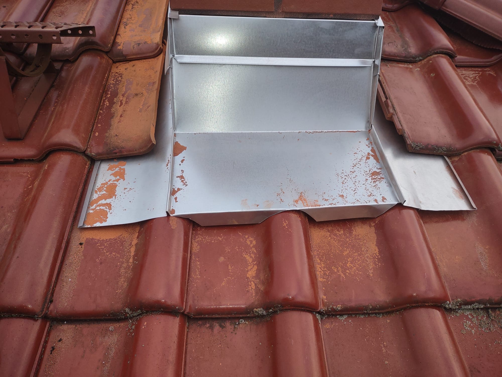 Szczegółowe ujęcie na nową, srebrną obróbkę blacharską komina na dachu z czerwonej dachówki ceramicznej. Widoczne ślady użytkowania na dachówce.