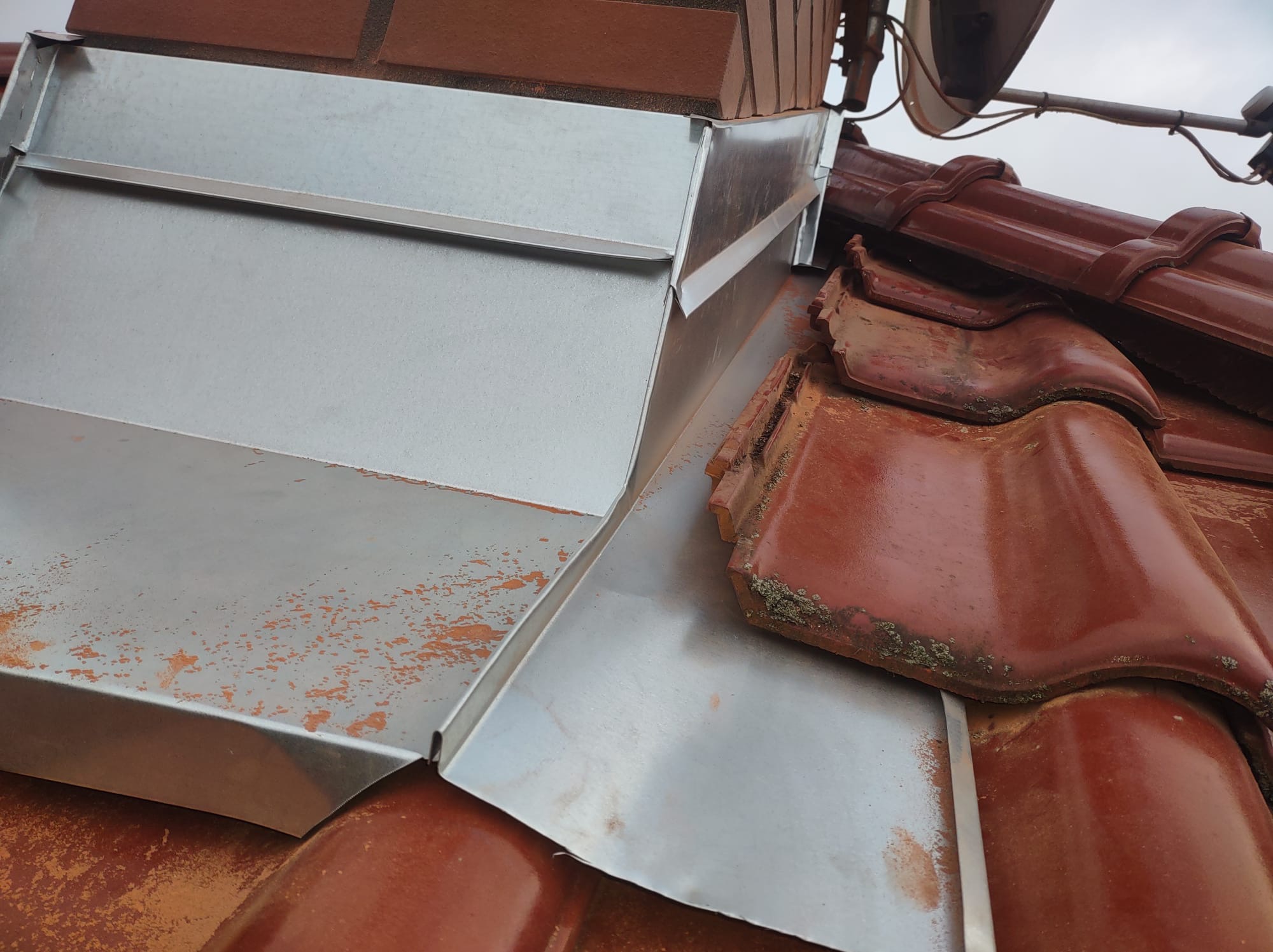 Szczegółowe ujęcie połączenia komina z dachem, z widocznymi obróbkami blacharskimi z blachy ocynkowanej i fragmentem ceramicznej dachówki w kolorze ceglastym.